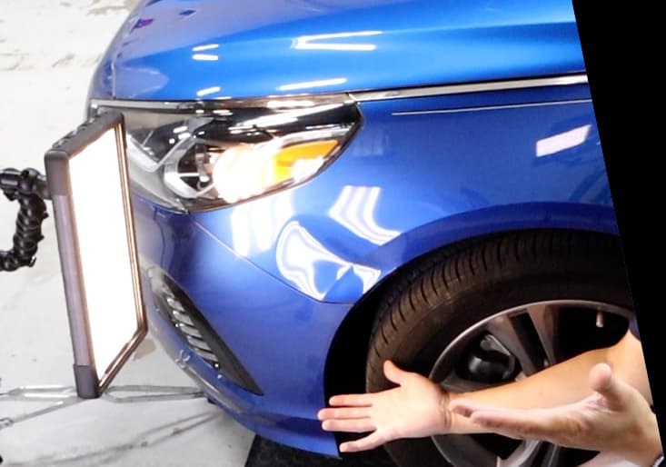 A person inspecting the front wheel and headlight of a blue car.